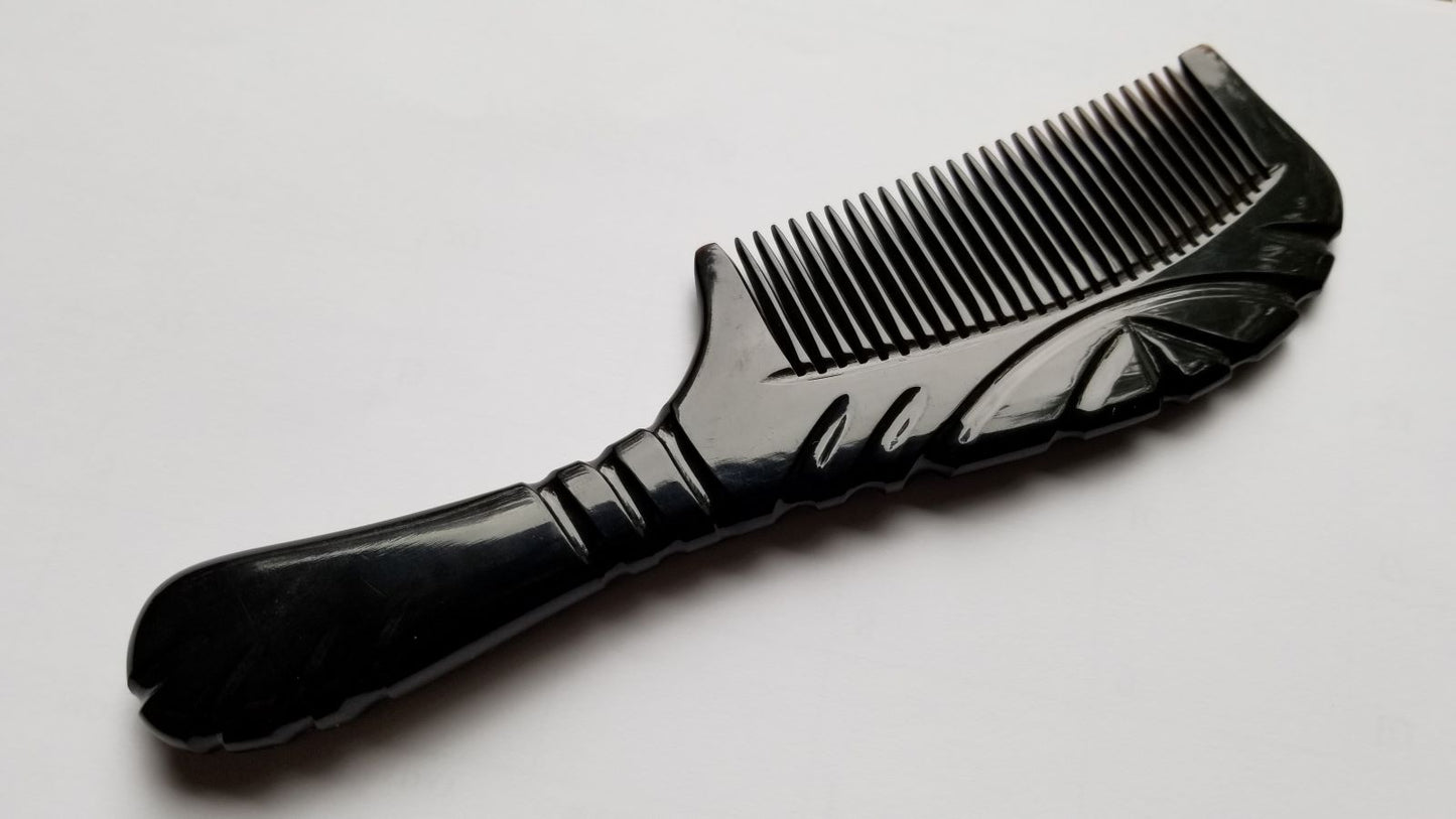 A black comb with a decorative handle and teeth. The handle features carved line designs. The comb is made of buffalo horn and is designed for hair grooming. The comb is approximately 7 inches long.