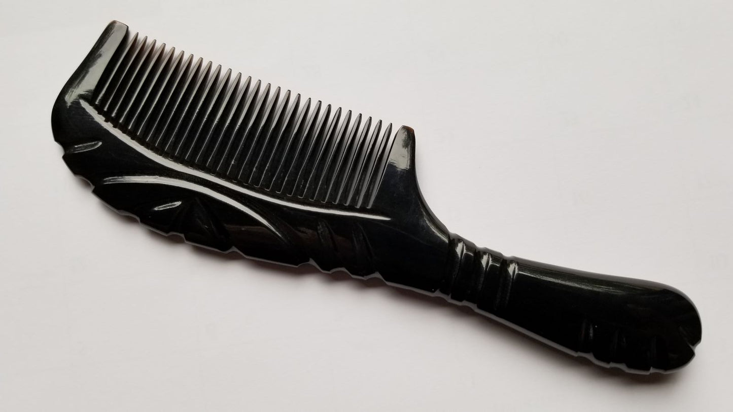 A black buffalo horn comb with a handle and fine teeth. The handle has a curved design with carved line details. The comb teeth are closely spaced.