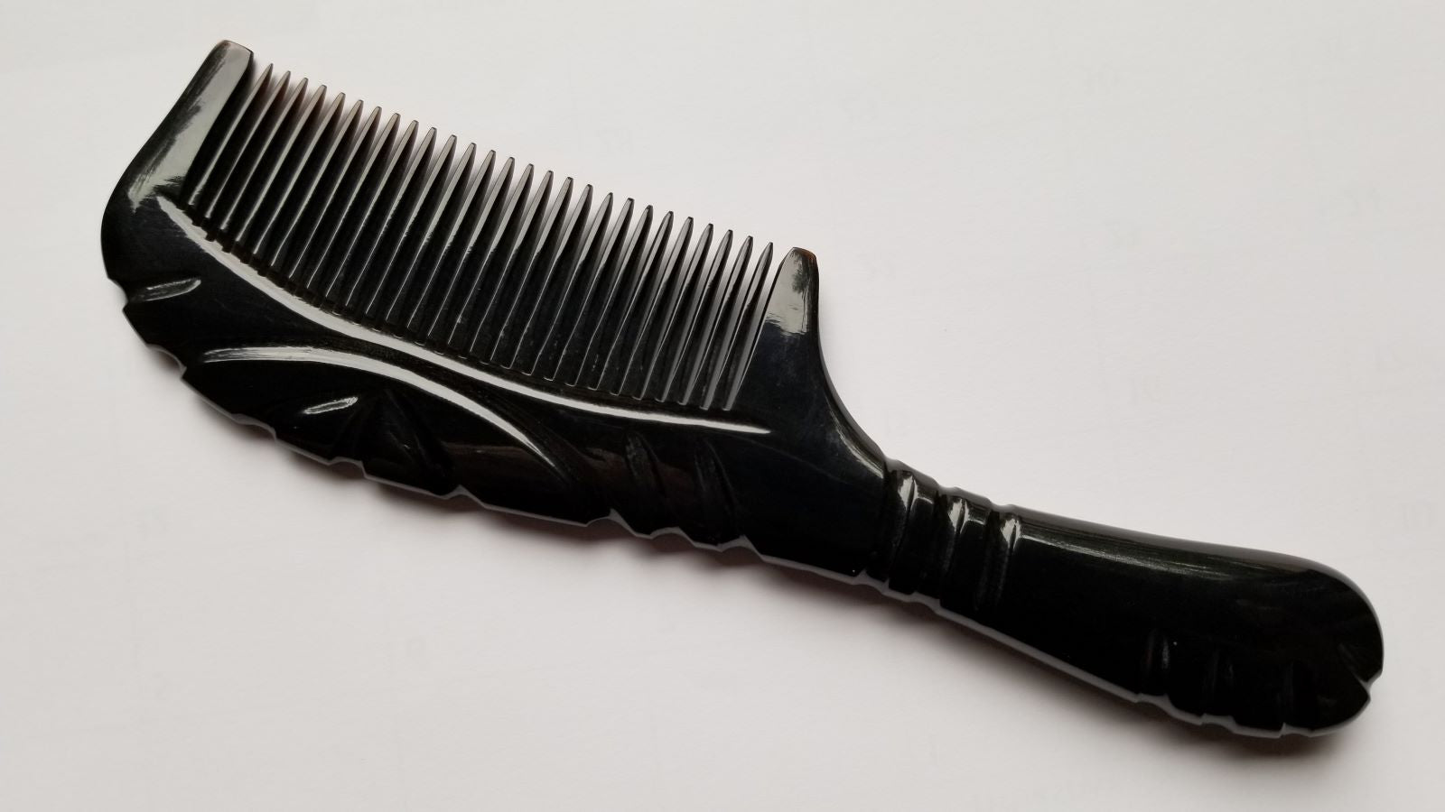 A black buffalo horn comb with a handle and fine teeth. The handle has a curved design with carved line details. The comb teeth are closely spaced.