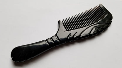 A black comb with a decorative handle and teeth. The handle features carved line designs. The comb is made of buffalo horn and is designed for hair grooming. The comb is approximately 7 inches long.