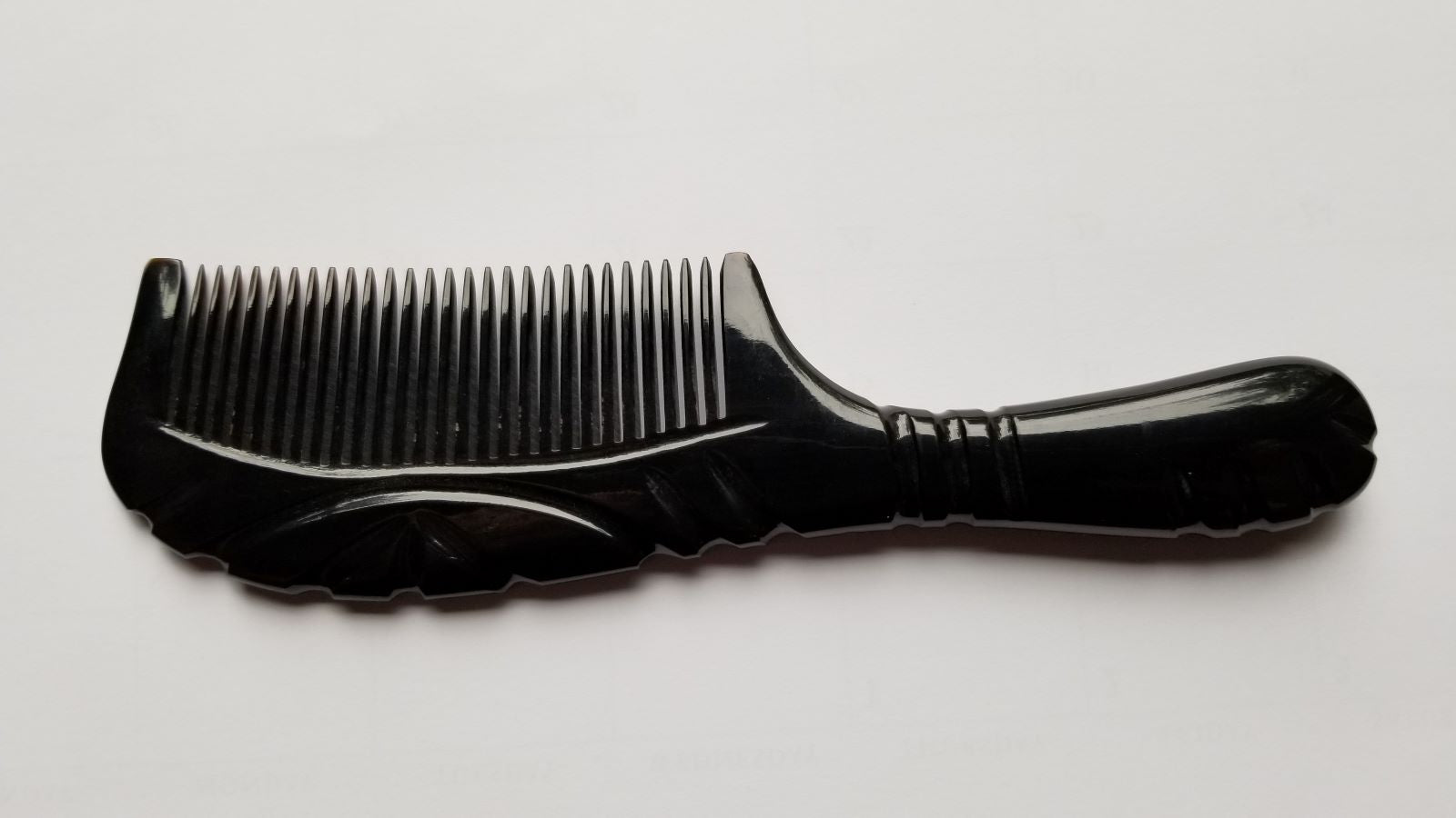 A black comb with a handle and teeth. The comb is made of a black smooth buffalo horn. The handle has a curved design and the teeth are evenly spaced.