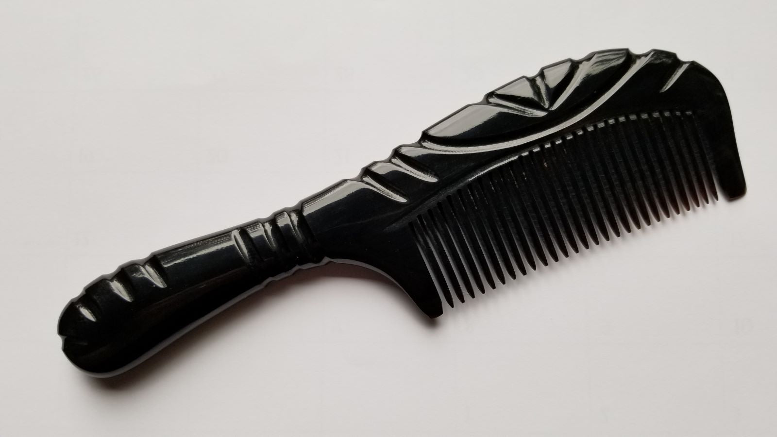 A black comb with a decorative handle and fine teeth. The comb has a glossy finish and a curved design. The handle features carved details, adding to its aesthetic appeal. This hair care accessory is designed for grooming.