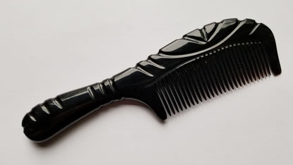 A black comb with a decorative handle and fine teeth. The comb has a glossy finish and a curved design. The handle features carved details, adding to its aesthetic appeal. This hair care accessory is designed for grooming.