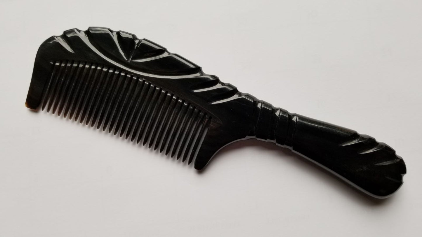 A black comb with a handle and fine teeth. The comb has a decorative, carved design on the handle and the top of the comb. The comb is made of black buffalo horn.
