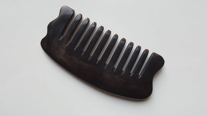 A near black buffalo horn comb with a curved handle and evenly spaced teeth. The comb is designed for hair grooming and styling.  The comb has curved handle and corners to use as massage plate.
