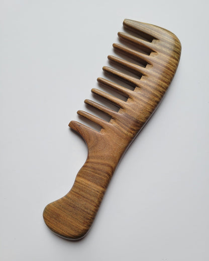 A wooden comb with wide teeth, featuring a curved handle for easy grip. The comb is made of light brown wood with a natural grain pattern. The teeth are evenly spaced and the overall design is simple and functional.