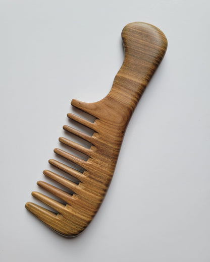 A handmade sandalwood comb with a extra wide-tooth design. The comb is made of a light brown wood with a curved handle and a series of evenly spaced teeth. The comb is designed for detangling and styling hair.