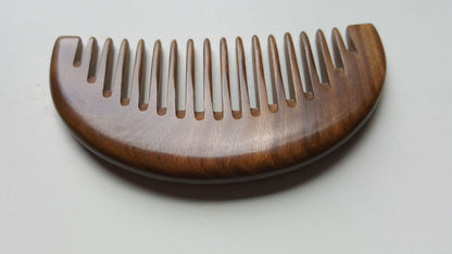 A brown wooden comb with a curved handle and wide teeth. The comb is made of natural sandalwood and has a smooth finish.  This comb has a beautiful wood grain pattern and is a one of kind piece.