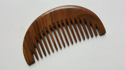 A dark brown wooden comb with a crescent shape and rounded teeth. It is made of green sandalwood and aromatic.