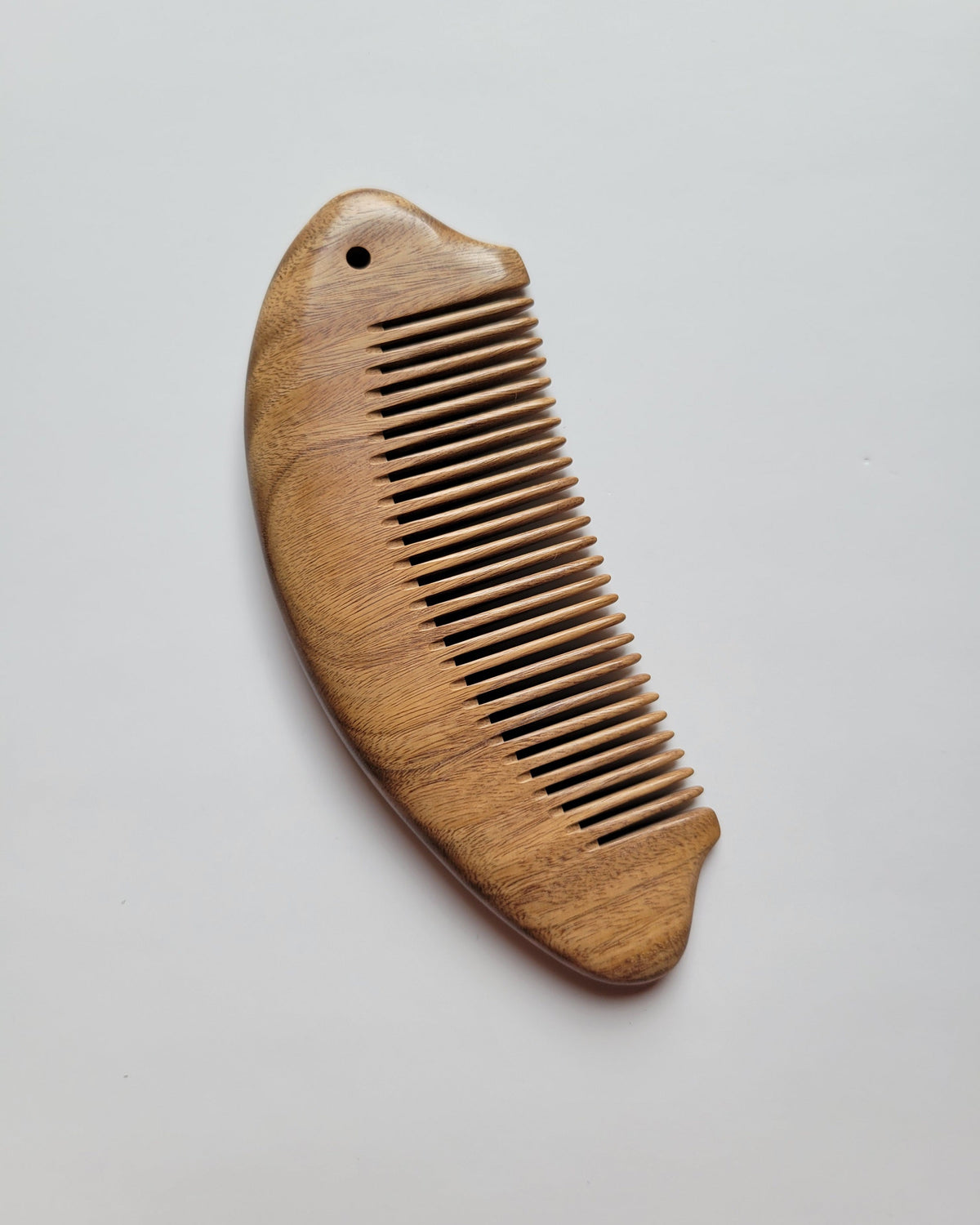 Hand carved green sandalwood fish comb, warm amber grain, arched fish body with tail fin and eye detail, fine tooth hair comb. A small hole is drilled into the handle.