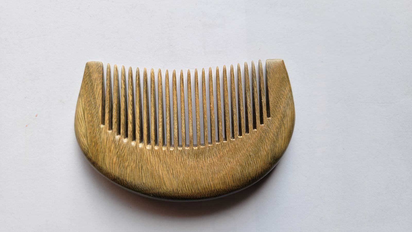 A wooden comb with a curved handle and closely spaced teeth. The comb is made of a light brown sandalwood with a visible grain pattern. The comb is designed for hair care and grooming.