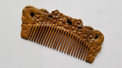 A wooden comb with intricate floral carvings. The comb's teeth are evenly spaced, and the handle features detailed relief carvings of flowers and leaves. The wood has a warm, natural tone, and the comb is set against a white background.