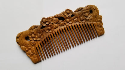 A wooden comb with intricate floral carvings. The comb is made of light brown wood and features detailed carvings of flowers and leaves along the top edge. The teeth of the comb are finely spaced. Comes with carrying case and gift for her. 