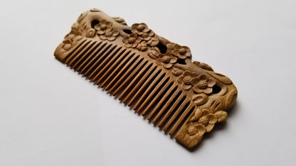 A hand carved sandalwoodcomb with intricate floral carvings. The comb is a warm brown color and features detailed carvings of flowers along the top edge. The teeth of the comb are evenly spaced and run the length of the comb. anti static and eco friendly gift. 