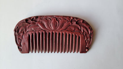 A red wooden comb with a carved floral design. The comb has a rounded shape and a row of teeth.