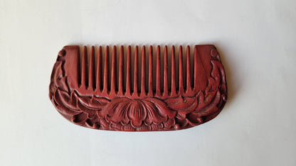 A red wooden comb with a carved floral design. The comb has a rounded shape and is made of a purple heart wood.