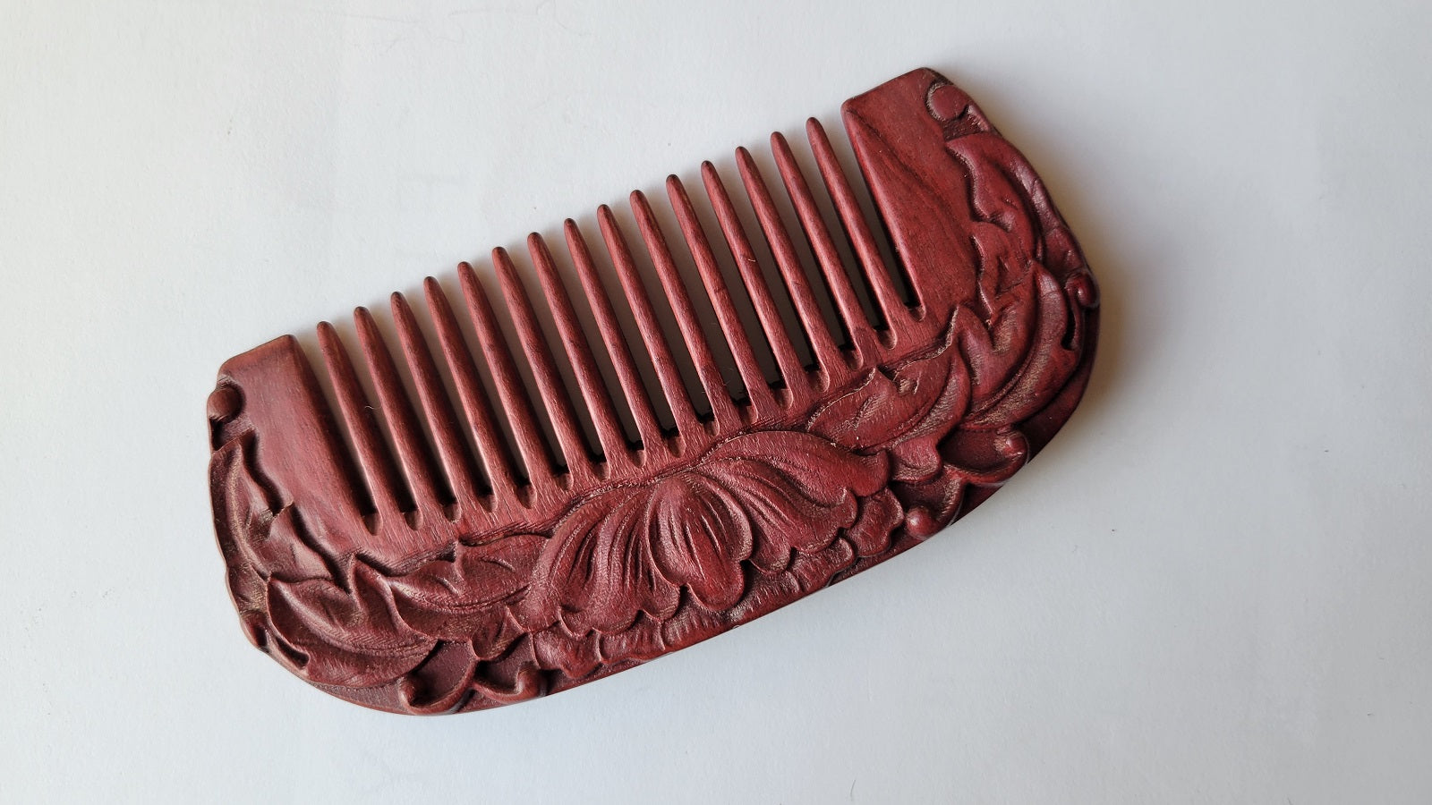 A purple heart wood comb with a carved lotus design on one side. The comb has a rounded shape and fine teeth.