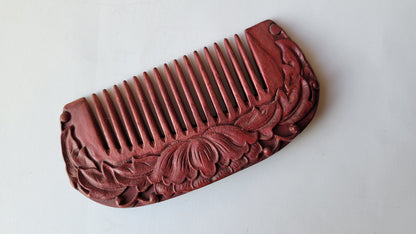 A purple heart wood comb with a carved lotus design on one side. The comb has a rounded shape and fine teeth.
