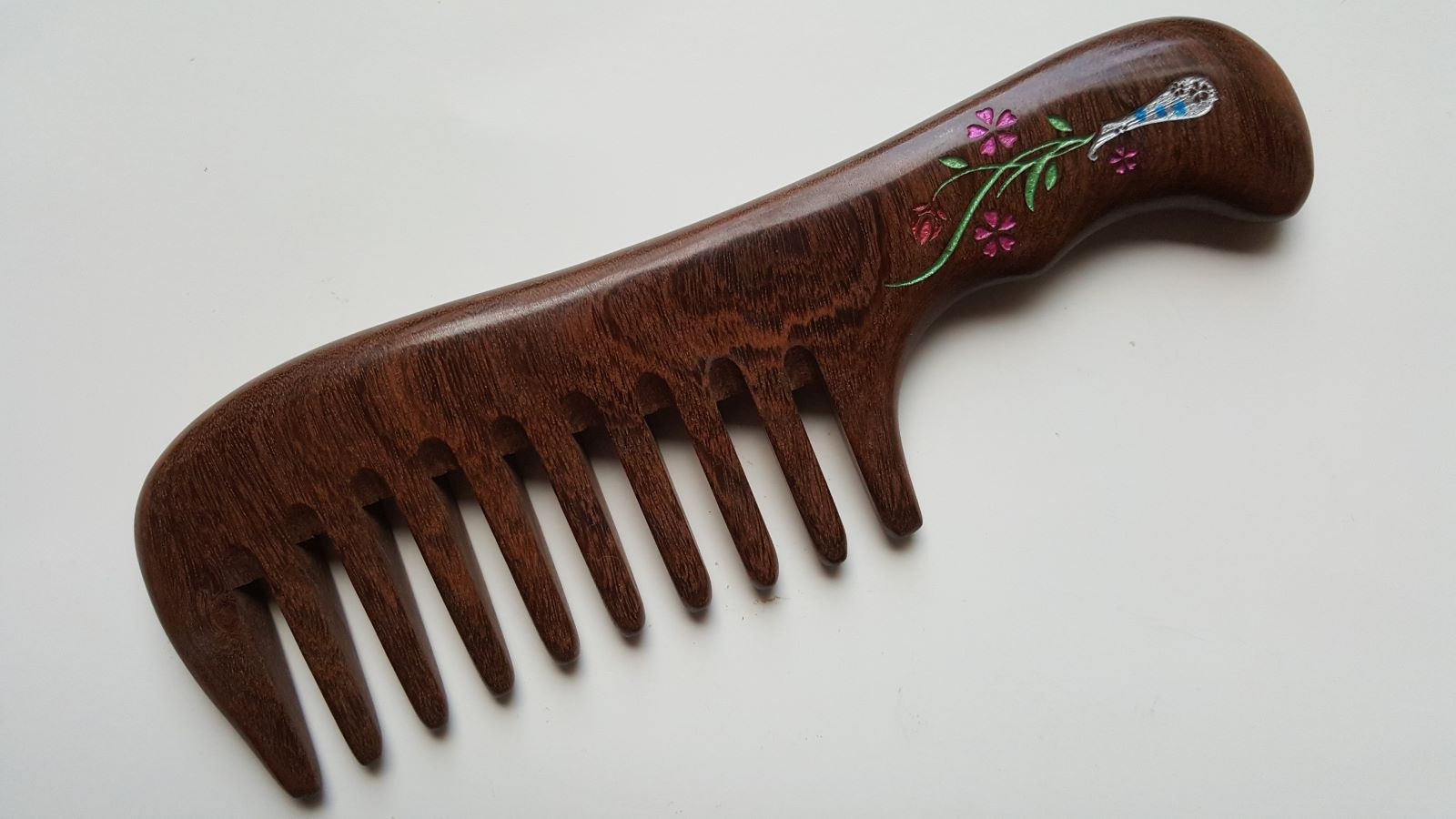 A brown Sucupira wood comb with a floral design slightly carved into the handle. The flowers are pink, green, and white. This wood comb has extra wide teeth and is good for curly, long, and thick hair. Gift for mom
