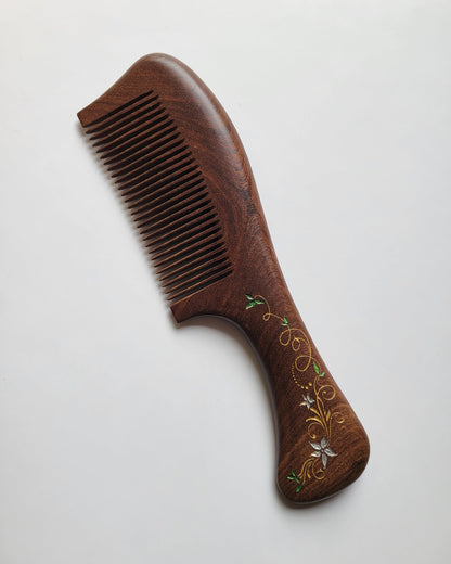 A brown wooden comb with a decorative ivy design on the handle. The comb has a curved shape and is made of natural Sucupira wood.