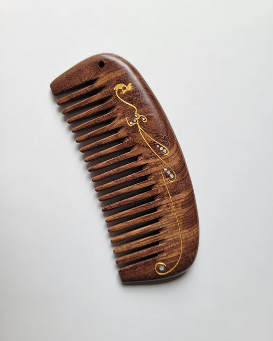 A wooden comb with a dark brown finish and a decorative gold design. The comb has evenly spaced teeth and a curved shape. The gold design features a flowing pattern with small, abstract accents.