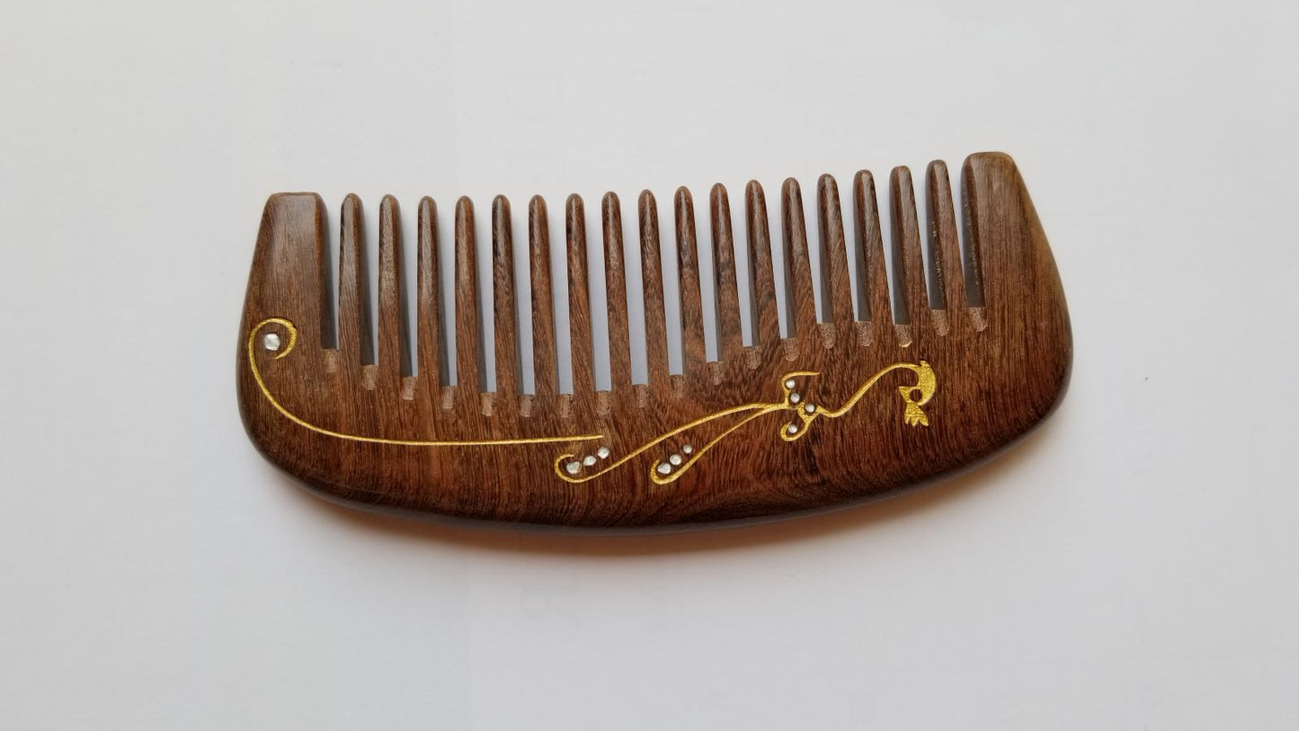 Dark brown Sucupira wood wide tooth comb, flat lay side angle showing curved shape and wood grain — Arakicrafts.  This comb features a gold-colored decorative design with small, clear embellishments. The design includes a bird-like shape and flowing lines.