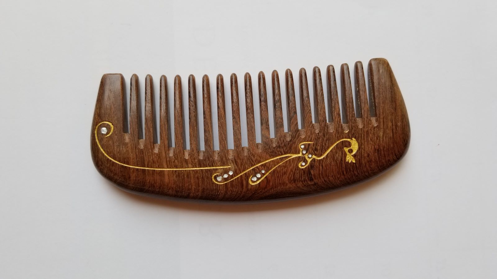 Dark brown Sucupira wood wide tooth comb, flat lay side angle showing curved shape and wood grain — Arakicrafts.  This comb features a gold-colored decorative design with small, clear embellishments. The design includes a bird-like shape and flowing lines.