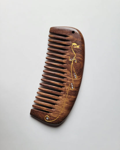 Dark brown Sucupira wood wide tooth comb, flat lay side angle showing curved shape and wood grain — Arakicrafts.  This comb features a gold-colored decorative design with small, clear embellishments. The design includes a bird-like shape and flowing lines.