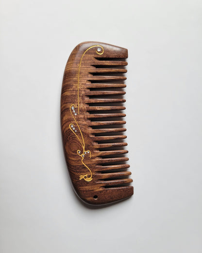Handcrafted wood comb with gold peacock motif, flat lay angled view of single-piece hardwood construction — Arakicrafts. The comb has closely spaced teeth and is designed for hair grooming.