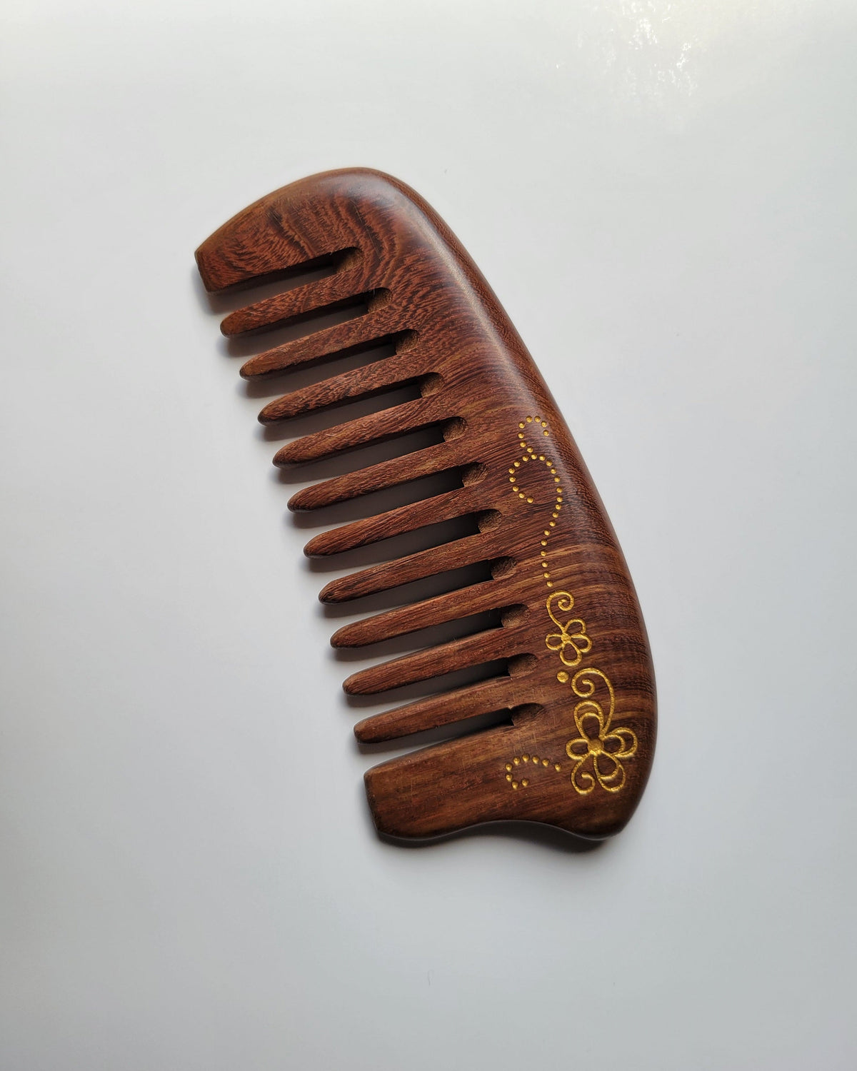 Gold floral scroll wood comb angled view, hand-painted four-petal flowers on deep reddish-brown Sucupira hardwood, wide tooth detangling comb