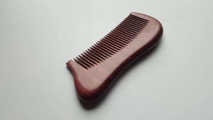 A wooden comb with a curved handle and closely spaced teeth. The comb is a rich, reddish-brown color and appears to be made of polished wood. The design is simple and functional, suitable for grooming.