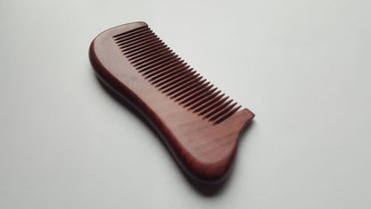 A wooden comb with a curved handle and closely spaced teeth. The comb is made of reddish-brown wood and is designed for grooming hair. The handle is ergonomically shaped for a comfortable grip.
