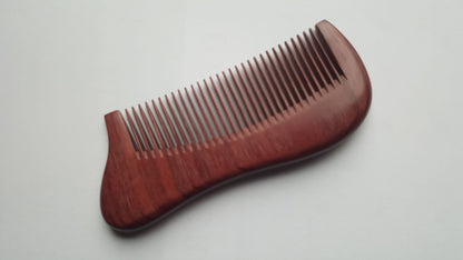 A wooden comb with a curved handle and closely spaced teeth. The comb is made of reddish-brown wood and is designed for grooming hair and beard. The handle is ergonomically shaped for a comfortable grip. A unique and special gift for men who appreciate fine quality products.