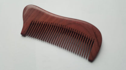 A wooden comb with a curved handle and fine teeth. The comb is made of a reddish-brown wood and has a smooth, polished finish. The teeth are evenly spaced and run the length of the comb. Eco-friendly and zero waste.