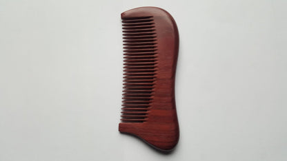 A wooden comb with a curved handle and fine teeth. The comb is made of a reddish-brown wood and has a smooth, polished finish. The teeth are evenly spaced and run the length of the comb. Eco-friendly and zero waste gift. 