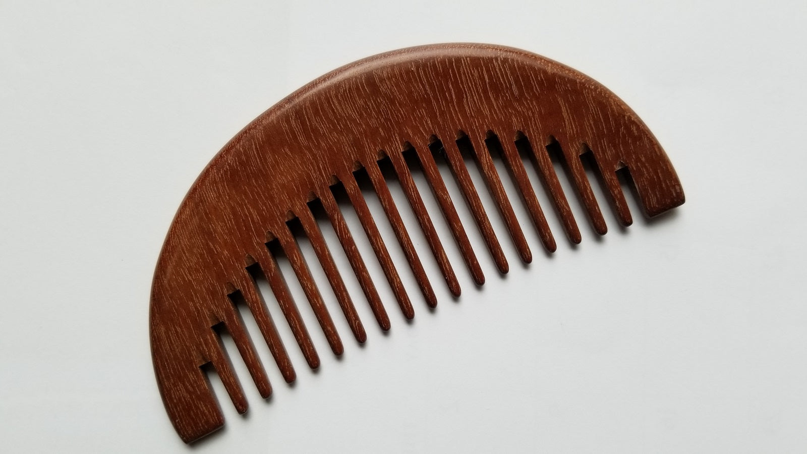 A wooden comb with a curved top and closely spaced teeth. The comb is made of a reddish-brown wood, and the grain is visible. The comb is designed for grooming hair and is a natural alternative to plastic combs.