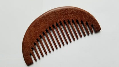 A wooden comb with a curved top and closely spaced teeth. The comb is made of a reddish-brown wood, and the grain is visible. The comb is designed for grooming hair and is a natural alternative to plastic combs.