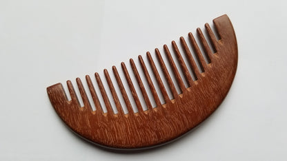 A wooden comb with a curved top and closely spaced teeth. The comb is made of a reddish-brown wood, and the grain is visible. The comb is designed for grooming hair and is a natural alternative to plastic combs. Perfect gift for eco conscious girl friends.