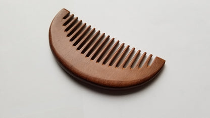 A wooden comb with a curved top and closely spaced teeth. The comb is made of a reddish-brown wood, and the grain is visible. The comb is designed for grooming hair and is a natural alternative to plastic combs. Perfect gift for mother's day gift!