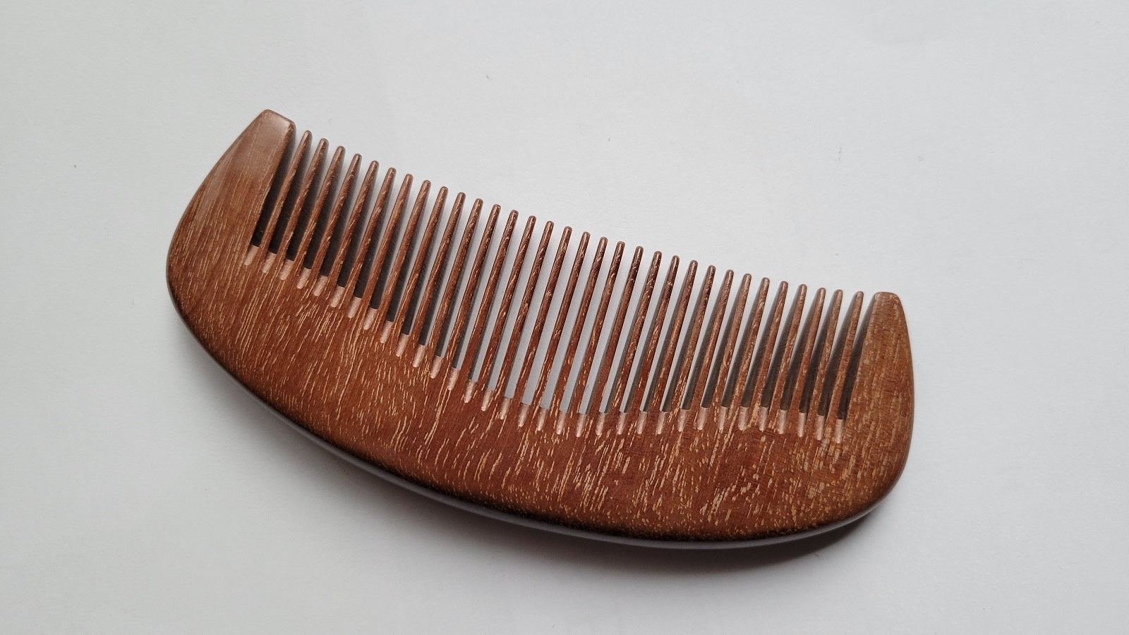 A wooden comb with a curved handle and closely spaced teeth. The comb is made of a reddish-brown wood and has a smooth, polished finish. The teeth are evenly spaced and extend from the handle.