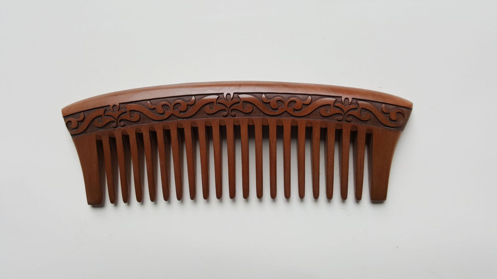 A brown wooden comb with a carved floral design. The comb has a curved handle and is made of natural peach wood.