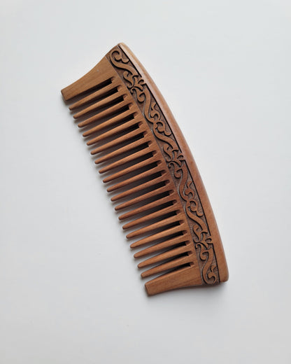 A brown wooden comb with a carved floral design. The comb has a curved handle and is made of natural peach wood.
