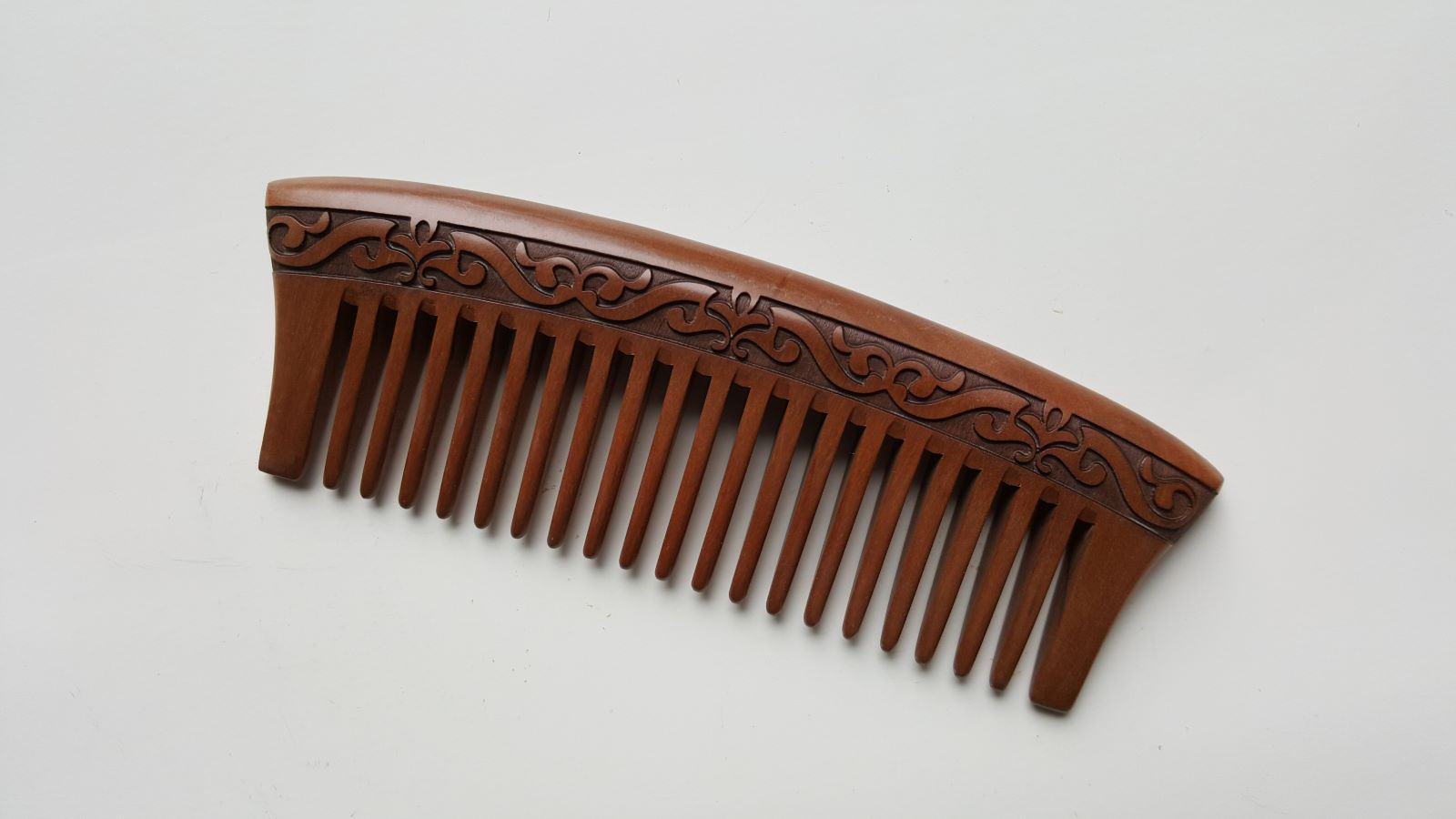 A brown wooden comb with a carved l design. The comb has a curved handle and evenly spaced wide teeth, good for daily hair grooming