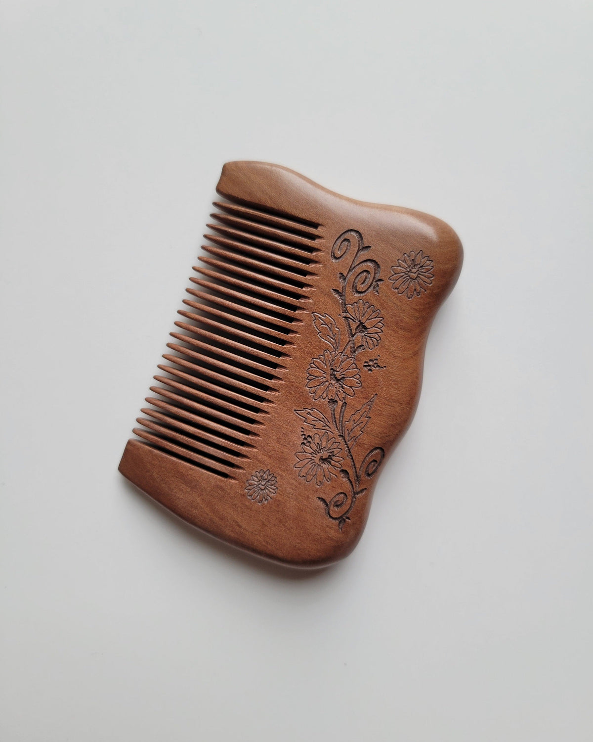 Angled view of a handcrafted floral comb showing delicate petal details and flowing scrollwork beside closely spaced wooden teeth.