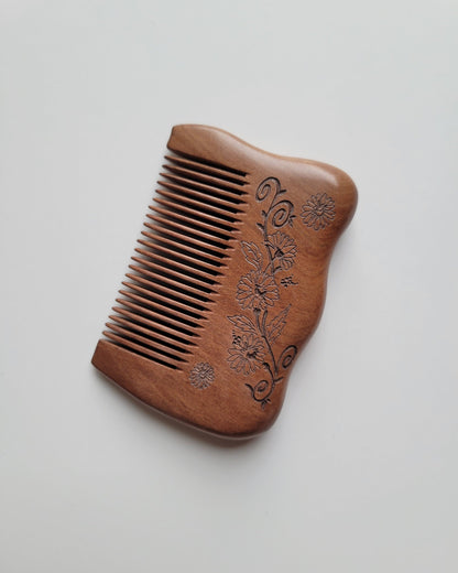 Angled view of a handcrafted floral comb showing delicate petal details and flowing scrollwork beside closely spaced wooden teeth.
