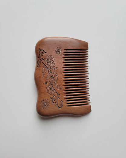 Overhead view of a small wooden comb with a gently curved spine, rounded corners, and floral carving, with teeth facing right.