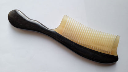 A black and beige comb with a curved handle. The comb has fine teeth and is made of a 100% natural wood and sheep horn.