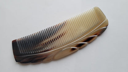 A brown and cream colored horn comb with a curved shape and a decorative design. The comb has a fine tooth design.