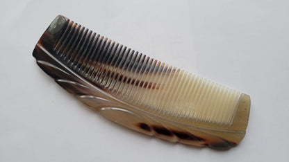 A brown and white tortoiseshell comb with a curved handle and a decorative design. The comb has fine teeth and is made of natural horn.
